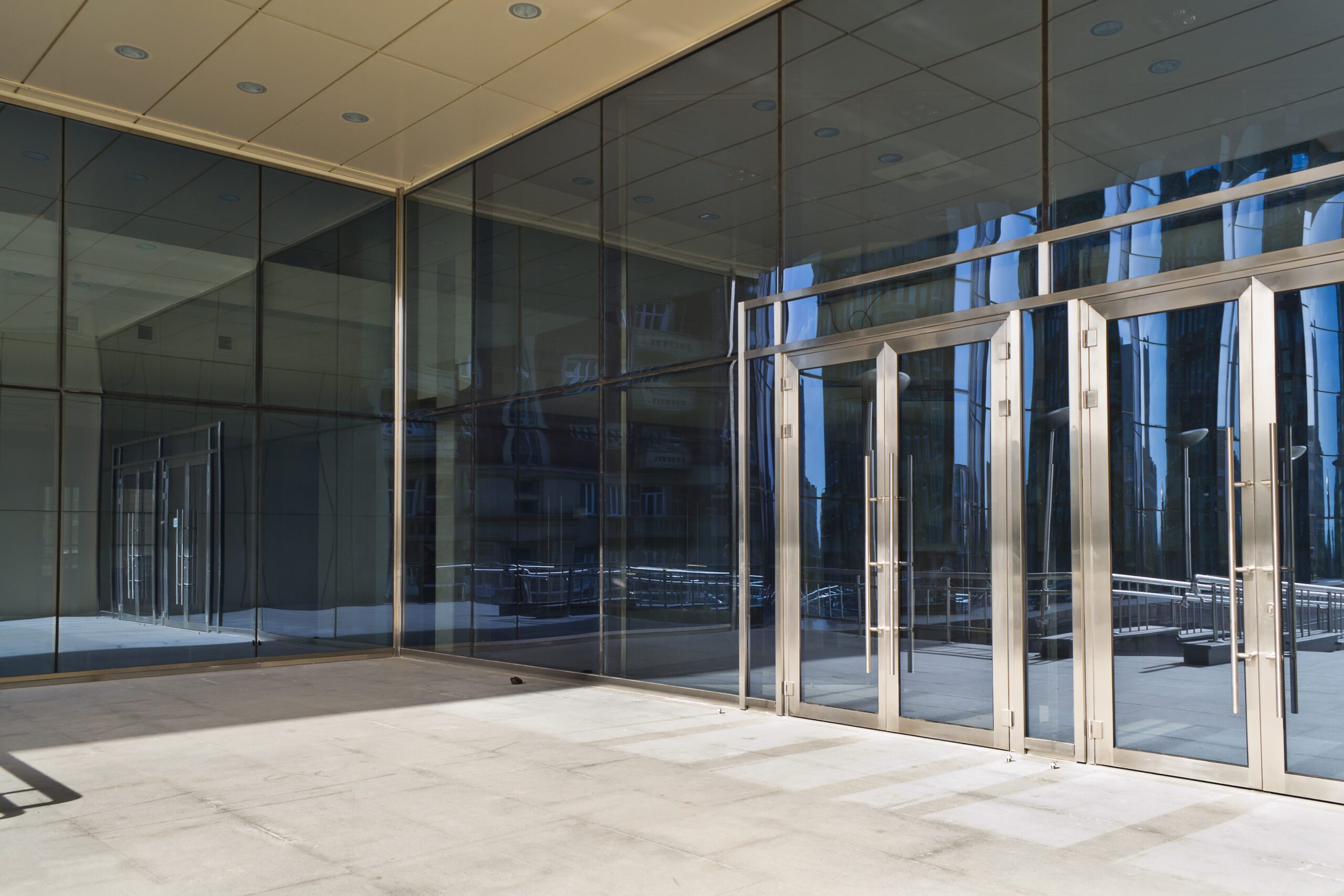 Exterior of a modern glass building with large reflective windows and multiple glass doors. The ground is concrete, and the scene is devoid of people.
